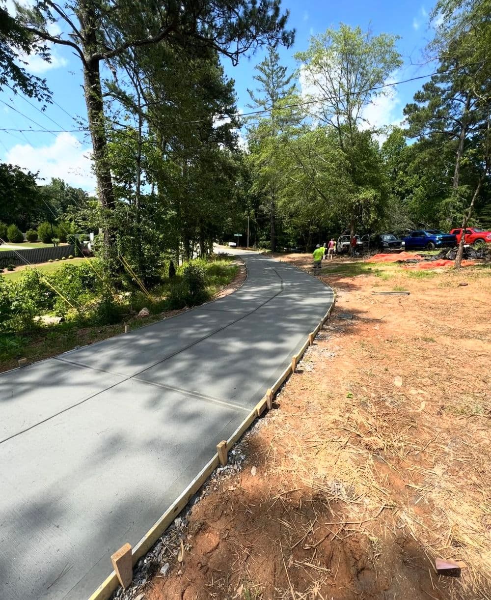 Newly poured concrete driveway winding through a landscaped area with trees and vehicles nearby.