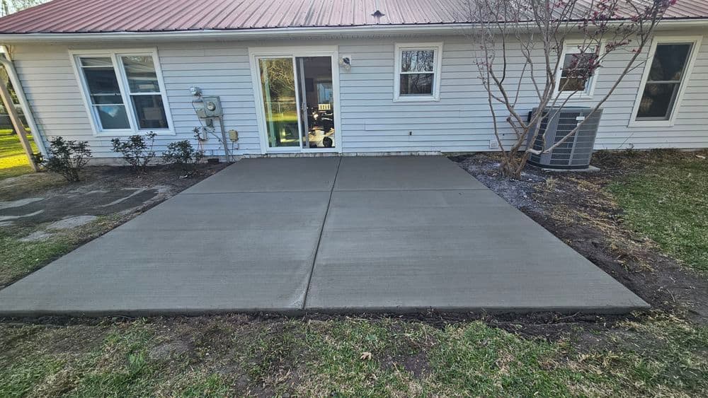 Newly poured concrete patio in backyard, adjacent to home with white siding and red roof.