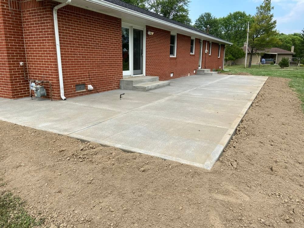 Concrete patio construction next to a brick house with yard space and trees in background.
