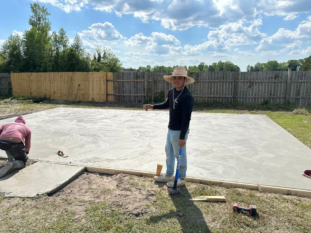 Construction worker pouring concrete on a slab in a backyard with cloudy blue sky.