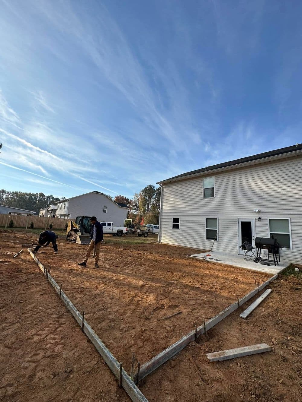 Construction workers preparing a home foundation in a residential backyard with clear skies.