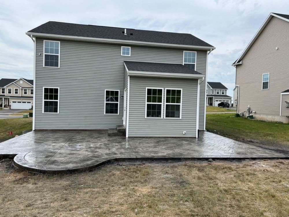 Newly installed stamped concrete patio at the back of a gray two-story house with green lawn.