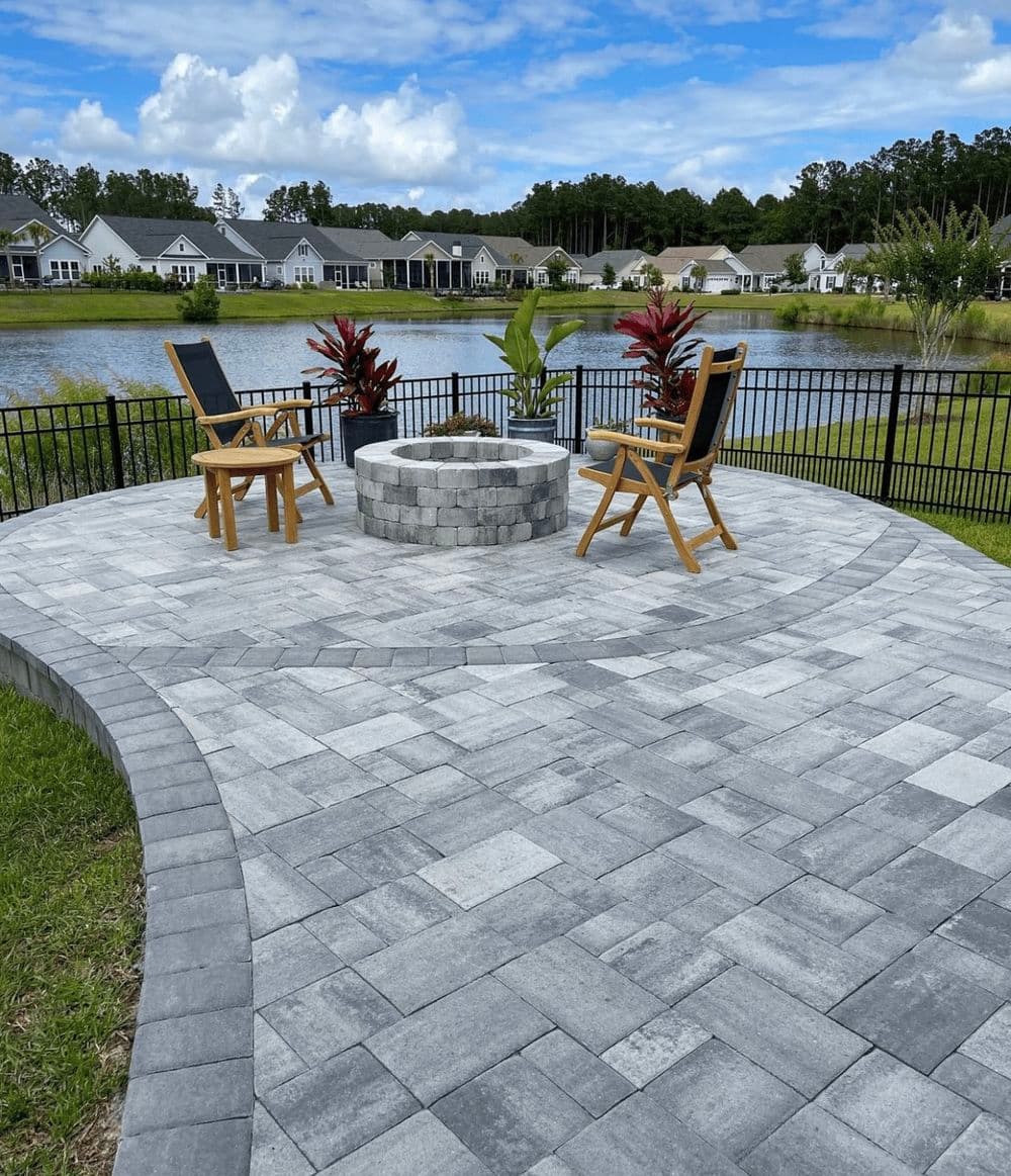 Outdoor patio with stone pavers, fire pit, and wooden chairs overlooking a serene lake.