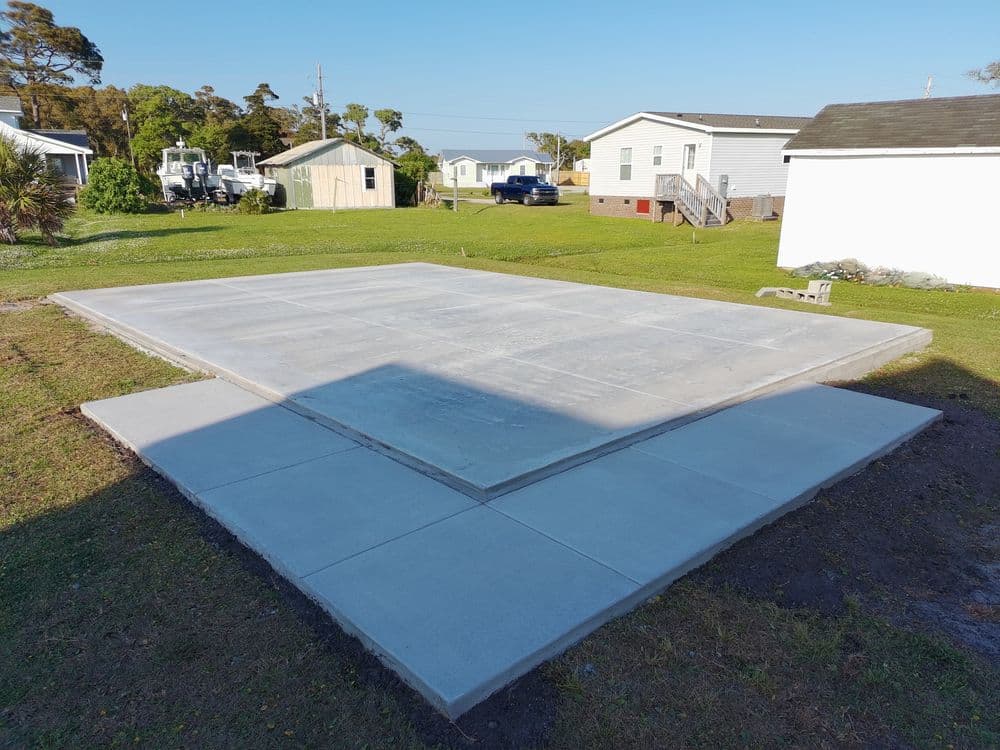 Concrete slab foundation ready for construction in a grassy yard with nearby structures.