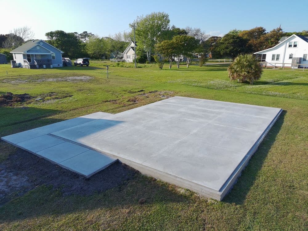 Concrete slab foundation on grassy land, surrounded by trees and houses. Ideal for construction.