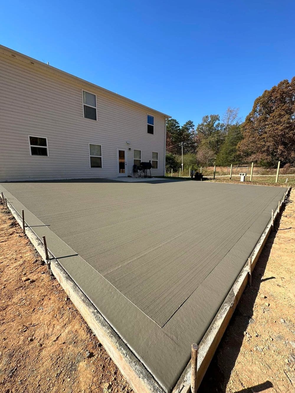Freshly poured concrete patio installation behind a house, clear blue sky above.