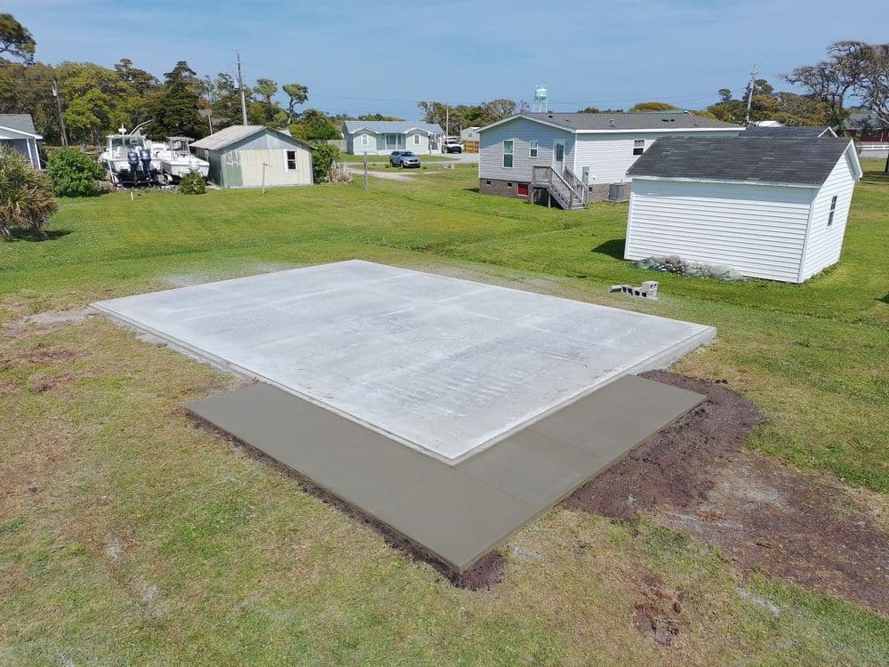 Concrete slab foundation on grassy lot, surrounded by houses and trees, under clear blue sky.