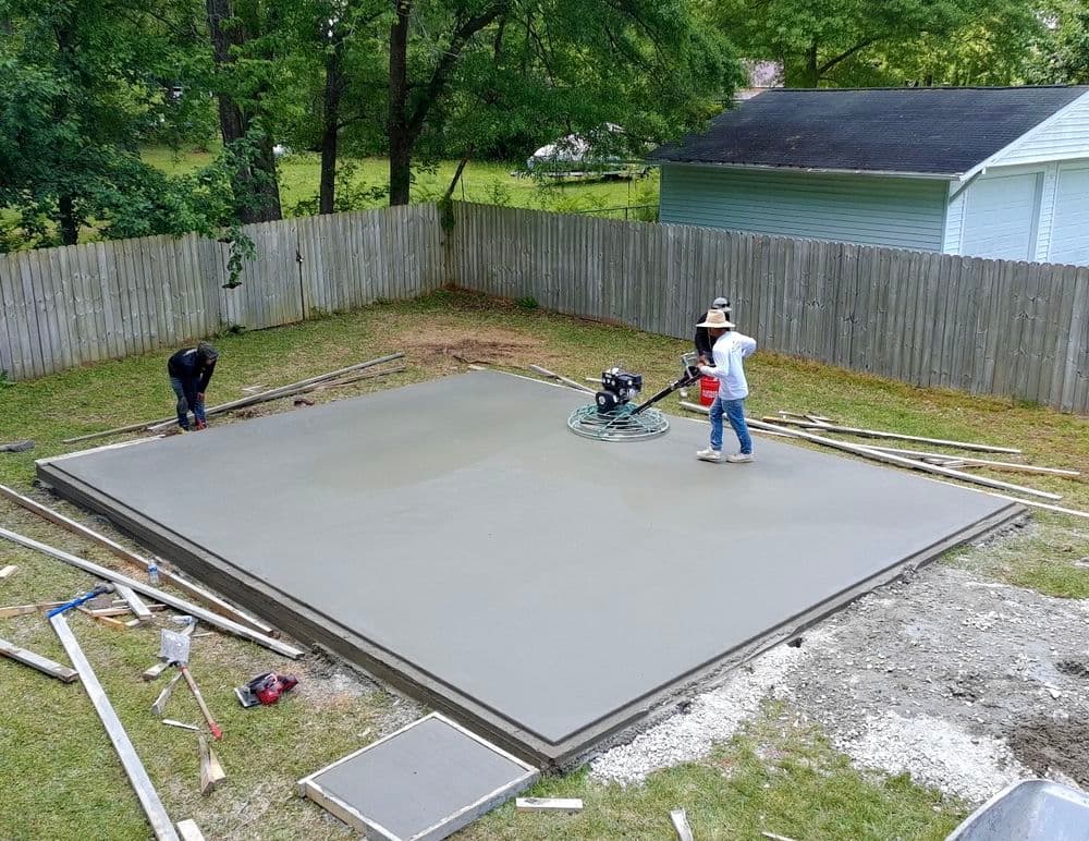 Workers pouring and finishing a concrete slab in a backyard under clear skies.