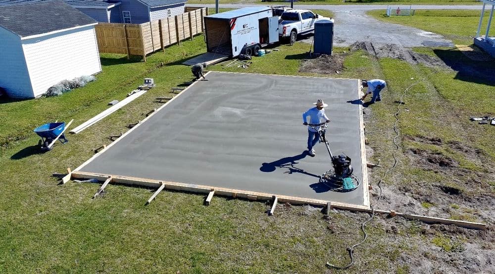 Workers leveling freshly poured concrete on a large outdoor slab with tools and equipment nearby.