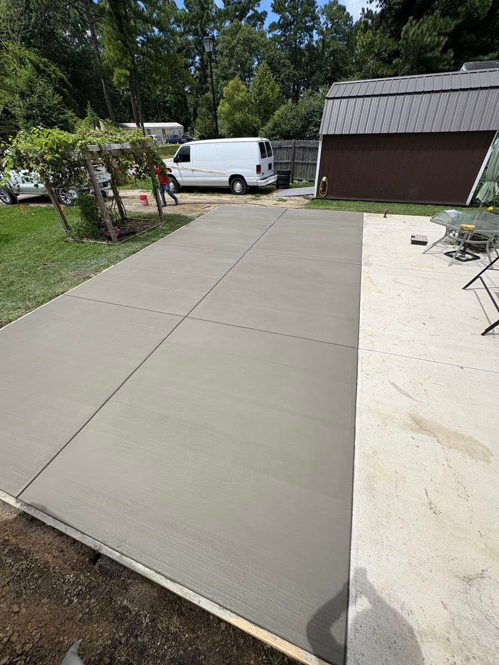 Freshly poured concrete slab in a backyard with green trees and a white van in the background.