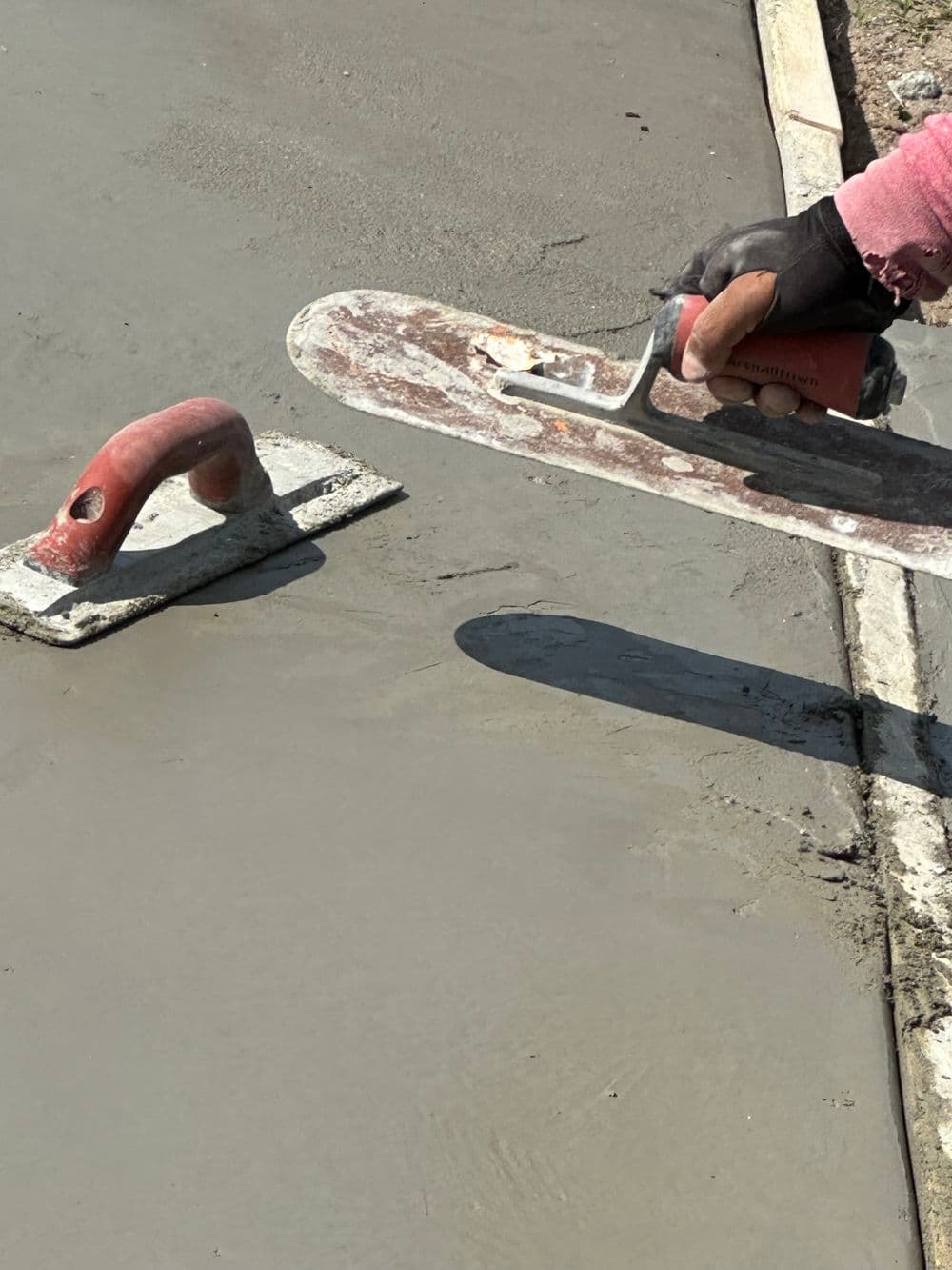 Worker applying concrete with a trowel next to a concrete float on a fresh slab.