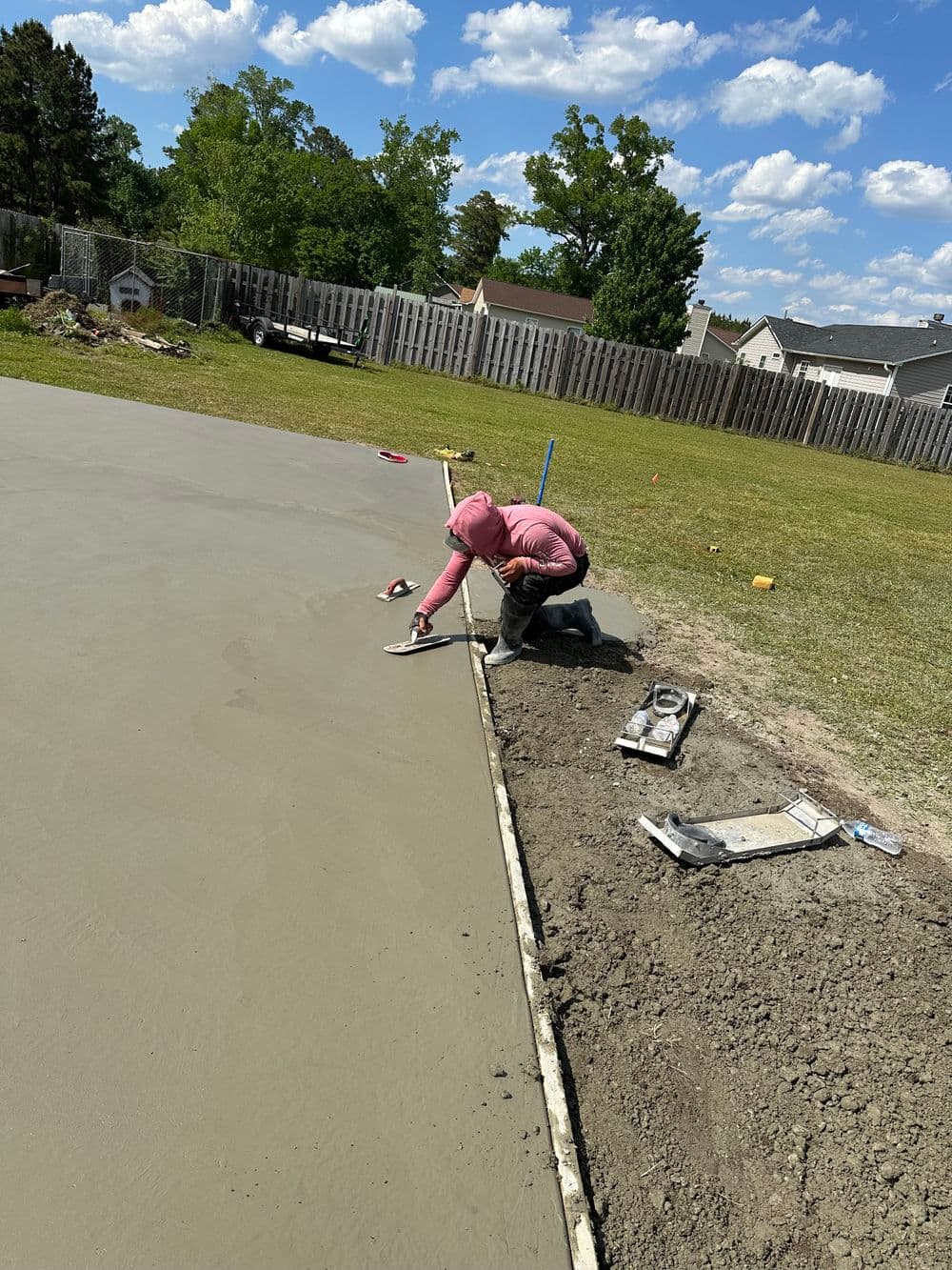 Person smoothing wet concrete on a sunny day, surrounded by grass and trees.