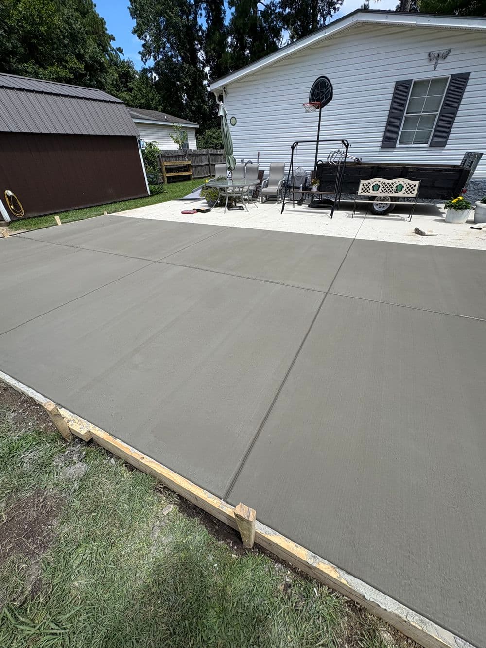 Newly poured concrete patio slab with wooden forms, surrounded by outdoor furniture and greenery.