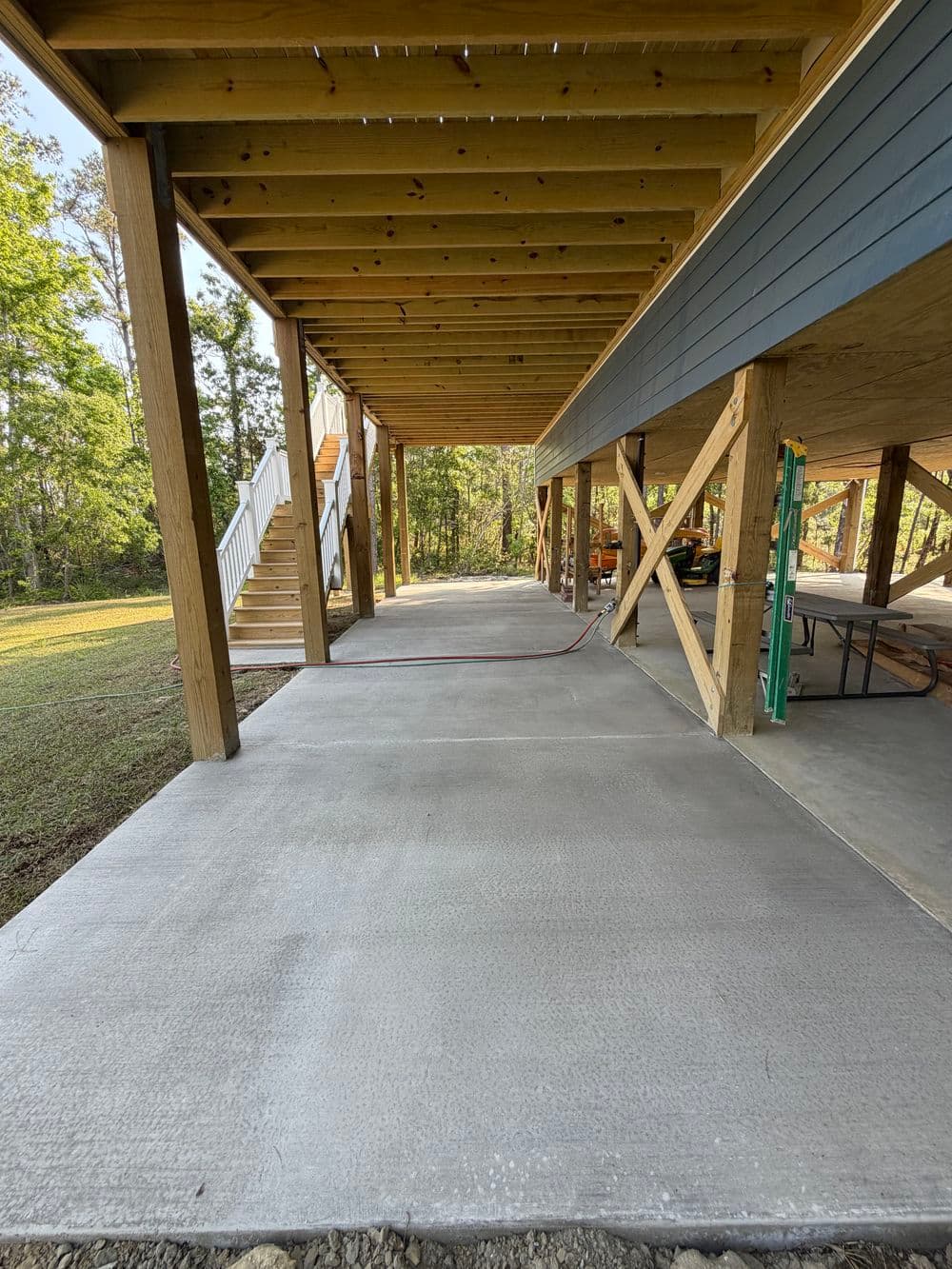 Newly poured concrete driveway under a raised house with wooden supports and stairs.