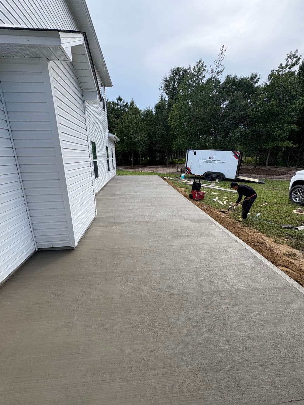 Concrete walkway installation beside a white house in a wooded area, with a worker in the background.