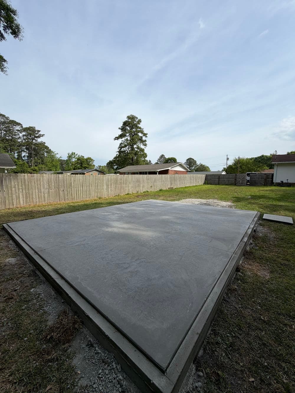 Newly poured concrete slab in a grassy yard, surrounded by wooden fence and trees.
