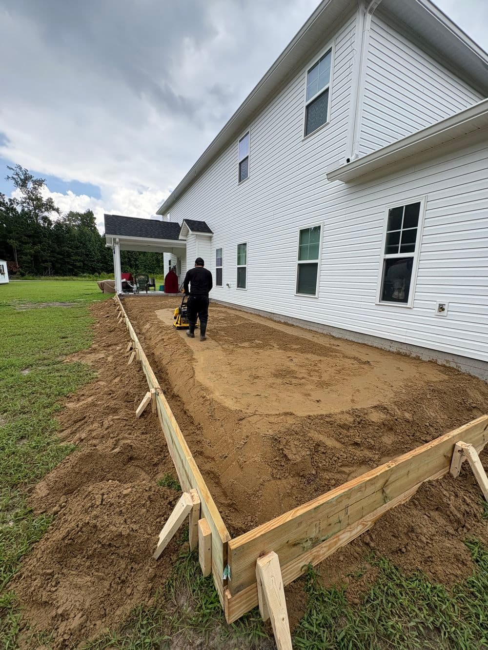Construction worker leveling a sand base for a patio beside a white house.