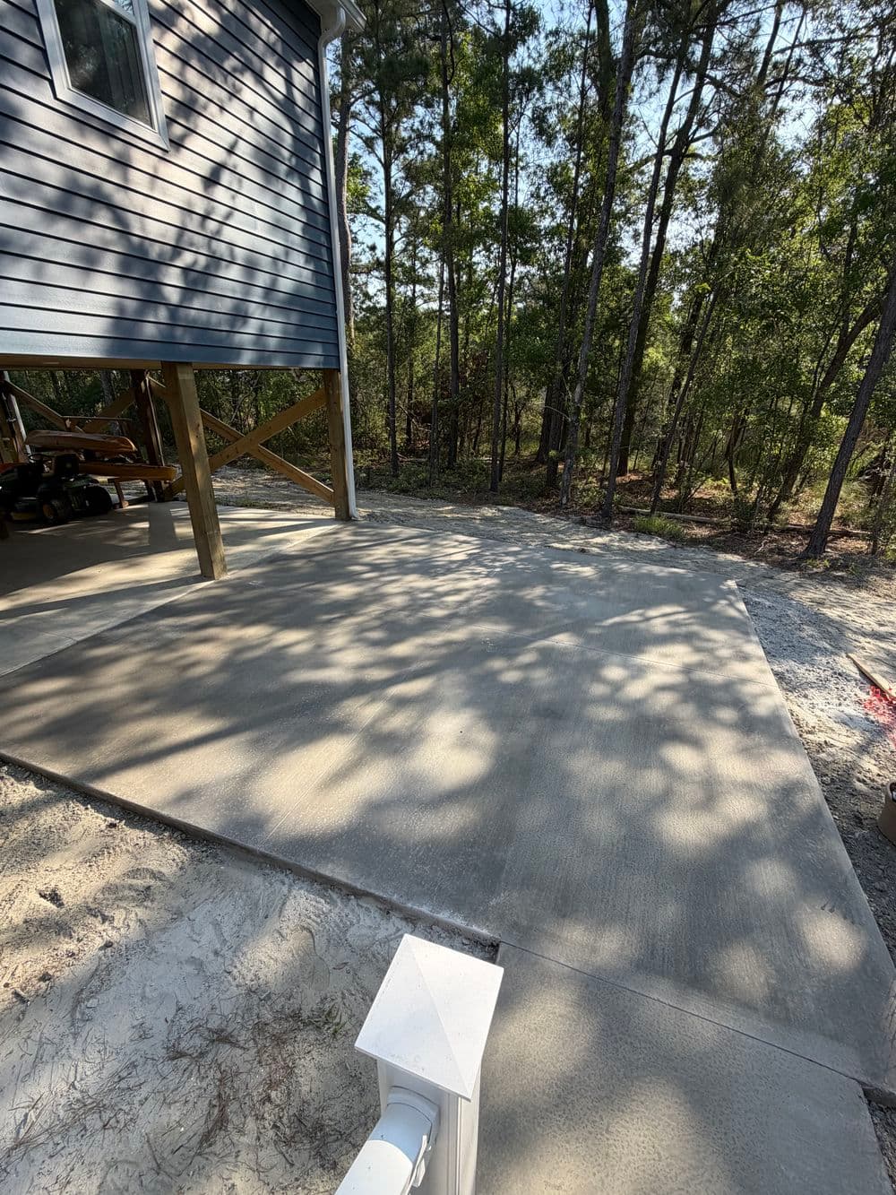 Freshly poured concrete patio beside a house in a wooded area with sunlight filtering through trees.