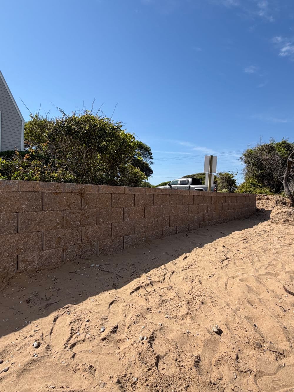 Sand-covered area near a stone wall, with shrubs and a blue sky in the background.