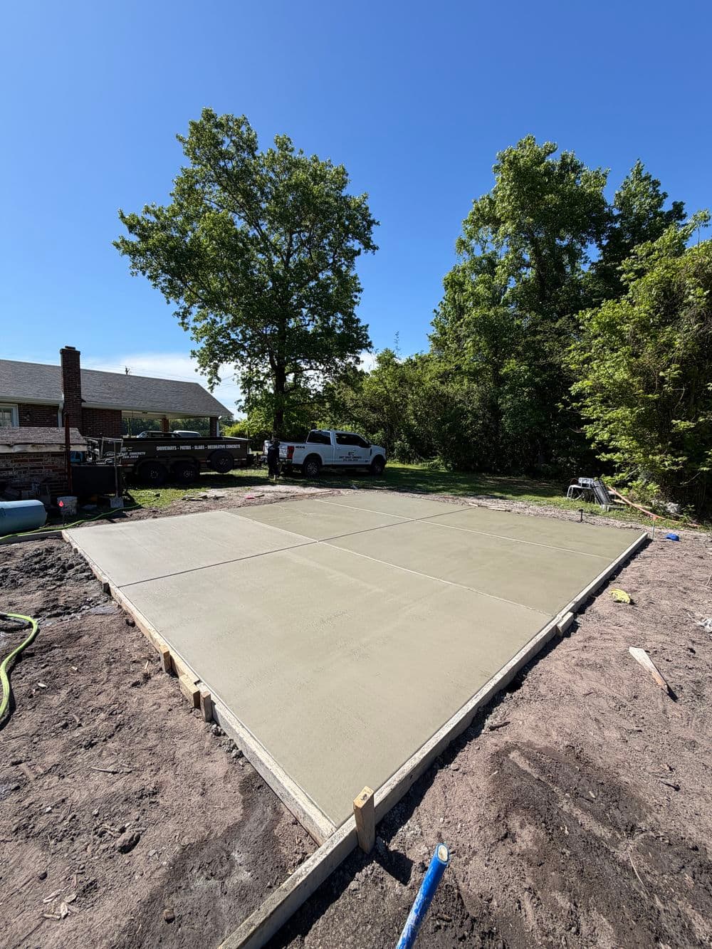 Freshly poured concrete slab in a sunny backyard surrounded by trees and a vehicle.