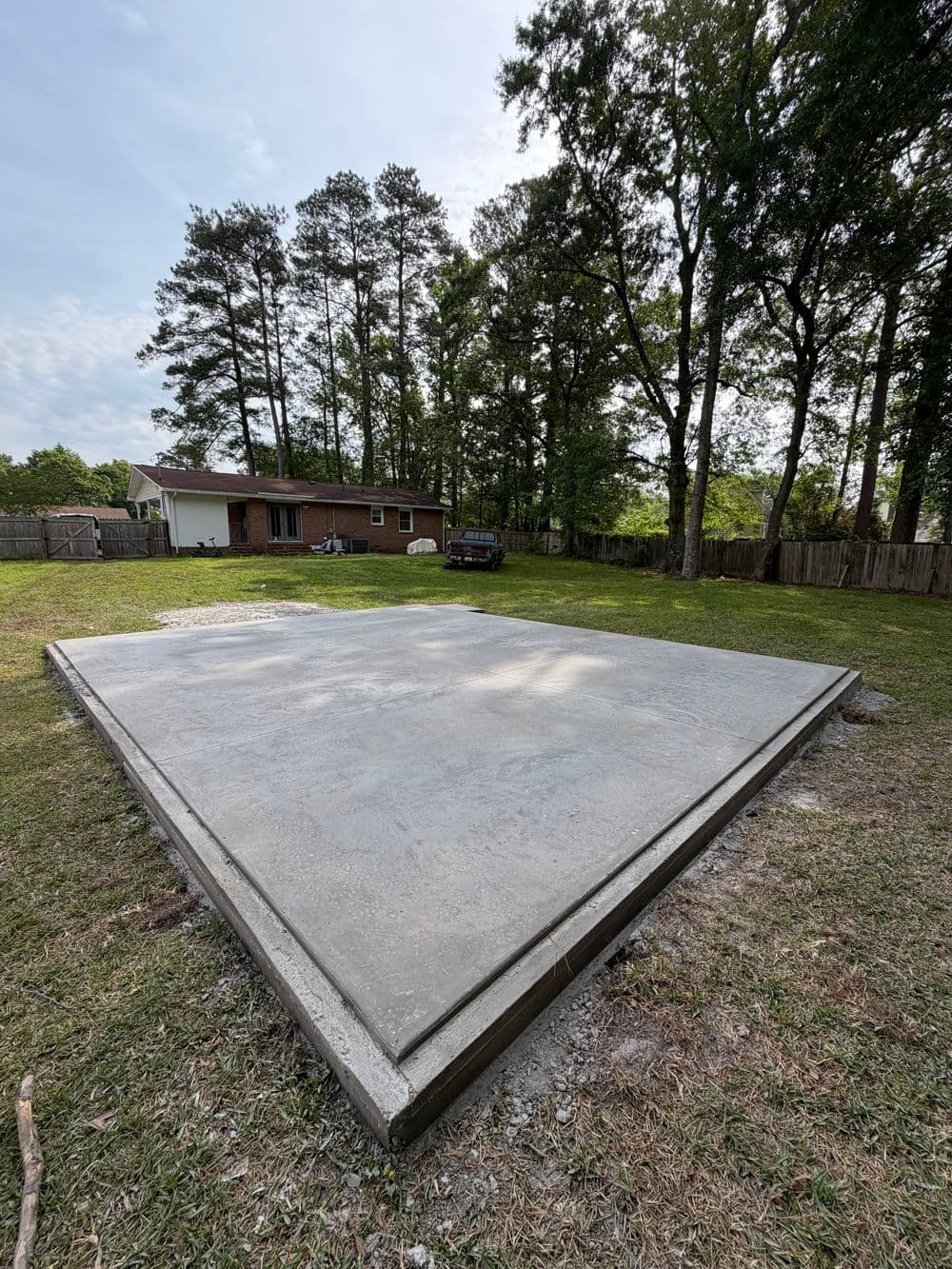 Concrete slab foundation in a grassy yard, surrounded by trees and a house in the background.