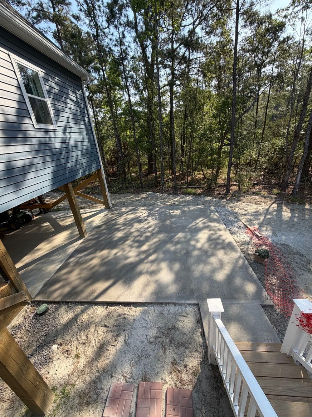 Newly poured concrete slab next to a house surrounded by trees and a gravel pathway.