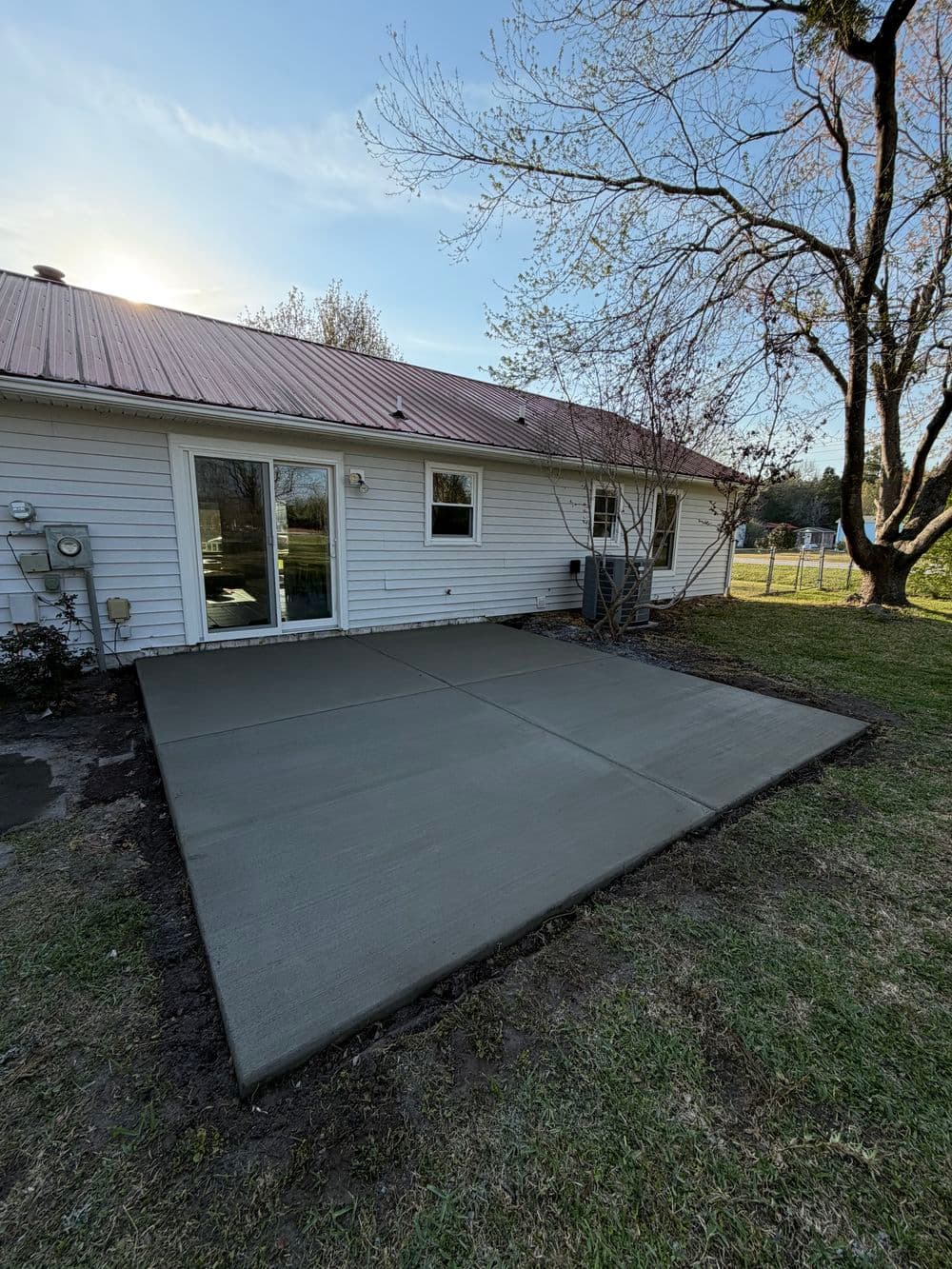 Newly poured concrete patio in backyard with white house and trees under blue sky.