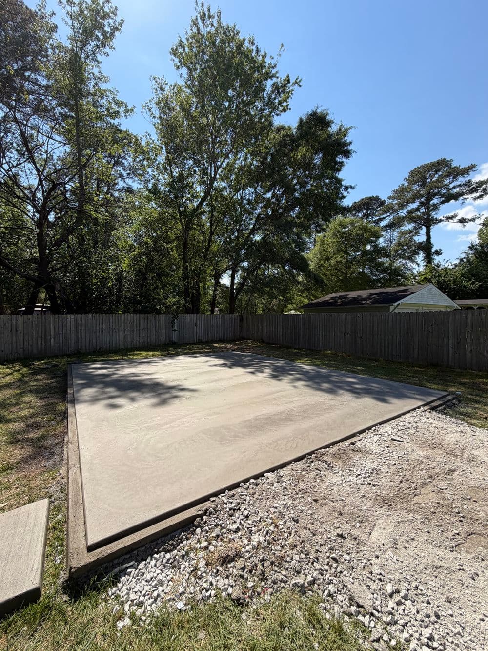Concrete slab foundation in a backyard, surrounded by trees and a wooden fence.