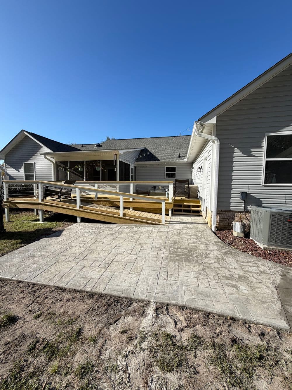 Accessible home with a wooden ramp, concrete patio, and landscaped yard under clear blue sky.